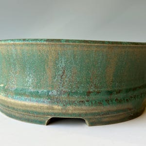 May include: A handcrafted, wide, oval-shaped ceramic bonsai pot. The pot features a textured surface with a blend of green and brown hues. The design includes a slightly flared rim and a series of horizontal ridges near the base, with small feet.