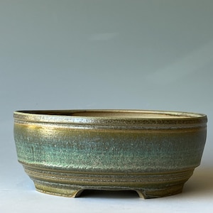 May include: A ceramic bonsai pot with a wide, oval shape. The pot features a textured, layered design with a gradient of colors, including green, gold, and brown. The pot has a low profile and is supported by small feet.