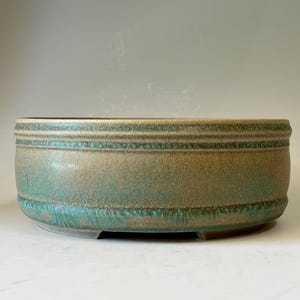 May include: A round ceramic bonsai pot with a tan base glaze and a textured, speckled teal green glaze overlay. The exterior features two horizontal bands of raised, decorative detailing around the upper and lower sections of the vessel.