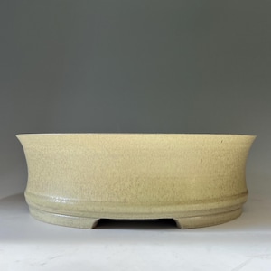 May include: A shallow, round ceramic bonsai pot with a pale yellow, speckled glaze. The vessel features a slightly flared rim, a smooth cylindrical body, and a raised base with small cutouts along the bottom edge, creating a stable foundation.