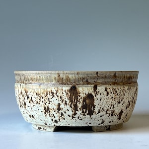 May include: A round ceramic bowl with a wide rim and a footed base. The exterior features a cream-colored glaze with a dense pattern of dark brown speckles and drips that are concentrated near the top rim and fade toward the bottom base.