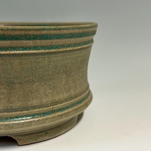 May include: A ceramic bowl with a wide, flared rim and a low, circular base. The exterior features a textured, matte beige finish with horizontal bands of vibrant teal glaze accenting the upper rim and the lower section near the base.