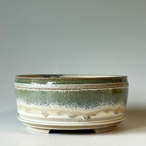 May include: A round ceramic bowl with a low profile and a wide opening. The exterior features a layered glaze with a deep green top section that transitions into a textured, speckled white and beige base with visible drip patterns.