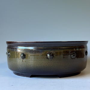 May include: A round, low-profile ceramic bowl with a dark brown and olive green glazed finish. The exterior features a series of evenly spaced, raised circular studs encircling the midsection of the vessel, which sits on a short, recessed base.