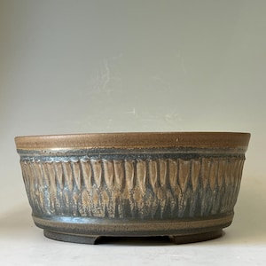 May include: A round ceramic bonsai pot featuring a textured exterior with vertical carved grooves. The glaze transitions from a smooth tan rim to a dark, mottled charcoal and brown finish on the lower body, resting on a short, raised circular base.