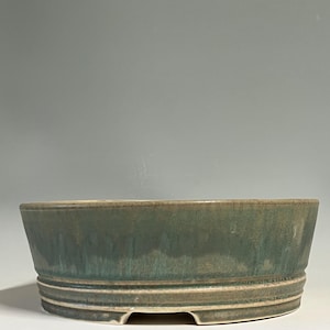 May include: A round ceramic bonsai pot featuring a muted green and blue glaze with a subtle drip effect. The exterior wall has a horizontal grooved band near the base, and the bottom edge includes small cutouts for drainage and airflow.