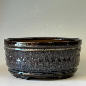 May include: A round, low-profile ceramic bowl with a dark brown and black glazed finish. The exterior features a textured band of vertical indentations around the center, while the interior has a smooth, glossy brown surface and a wide rim.