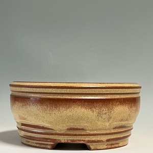 May include: A round ceramic bonsai pot featuring a two-tone glaze with a deep reddish-brown base color transitioning into a textured, sandy beige finish. The vessel has a wide, open top, a slightly tapered body, and a raised footed base.
