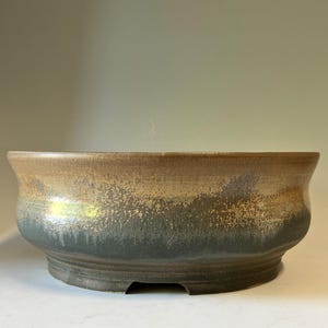 May include: A round ceramic bowl with a wide, flared rim. The exterior features a two-tone glaze, with a smooth, light tan upper section transitioning into a textured, dark charcoal gray base. The bowl has a thick, sturdy footed bottom.