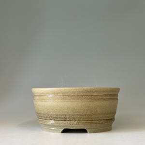 May include: A round ceramic bonsai pot with a tapered base and a wide, open top. The exterior features a mottled, earthy beige and tan glaze with subtle horizontal ridges circling the upper rim and a small cutout notch at the bottom edge.