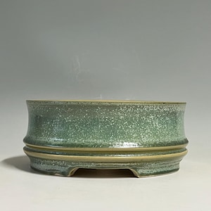 May include: A round ceramic bonsai pot with a speckled sage green glaze. The exterior features a smooth, slightly flared upper rim and a distinct horizontal recessed band above the base, which has small cutouts for drainage and airflow.