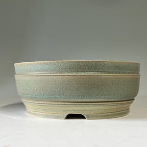 May include: A round ceramic bonsai pot with a speckled sage green and teal glaze. The pot features a wide, slightly flared rim, a smooth mid-section, and a raised base with a small drainage notch visible at the bottom edge of the vessel.
