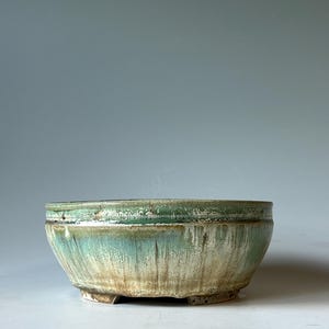 May include: A round ceramic bowl with a wide opening and a short, footed base. The exterior features a reactive glaze with blended shades of sage green, cream, and brown, displaying a vertical drip pattern that transitions to a darker rim.