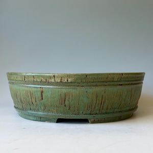 May include: A round, shallow ceramic bonsai pot with a green glaze featuring vertical brown drip patterns. The exterior has a raised horizontal band near the rim and a footed base with small cutouts along the bottom edge.