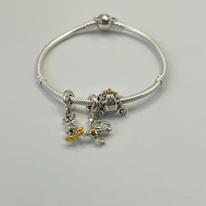 Puede incluir: Pulsera de plata con tres abalorios. Los abalorios incluyen un carruaje, una figura con pajarita dorada y un abalorio con un acento dorado. El abalorio del carruaje tiene el texto "Disney 100".
