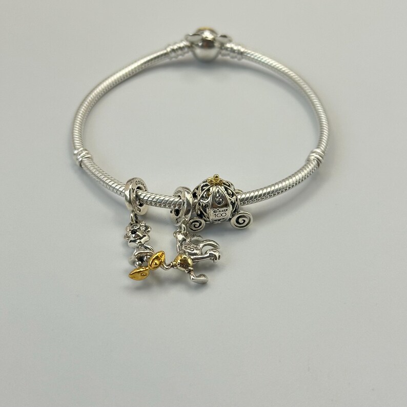 Puede incluir: Pulsera de plata con tres abalorios. Los abalorios incluyen un carruaje, una figura con pajarita dorada y un abalorio con un acento dorado. El abalorio del carruaje tiene el texto "Disney 100".
