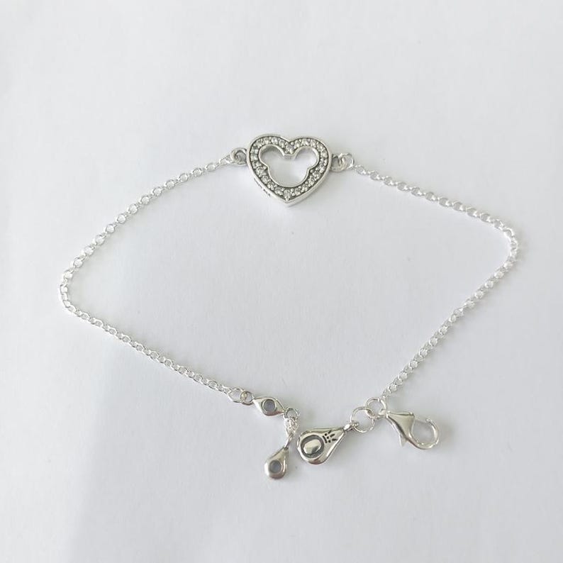 Puede incluir: Una pulsera plateada con un dije en forma de coraz&oacute;n con la silueta de Mickey Mouse. El dije est&aacute; incrustado con peque&ntilde;as piedras transparentes. La pulsera tiene una cadena delicada y un cierre de mosquet&oacute;n.