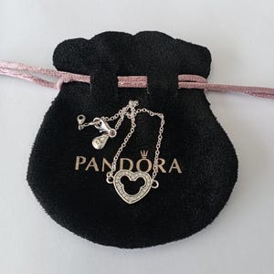 Puede incluir: Un collar de plata con un colgante en forma de coraz&oacute;n con un dise&ntilde;o de Mickey Mouse, engastado con peque&ntilde;as piedras transparentes. El collar se muestra en una bolsa de terciopelo negro con el nombre de la marca "PANDORA" impreso en blanco.