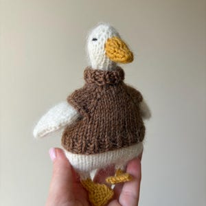 May include: Hand-knitted duck toy with a white head, wings, and body, a yellow beak and feet, and a brown sweater. The duck is a soft, plush toy, perfect for children or as a decorative item.