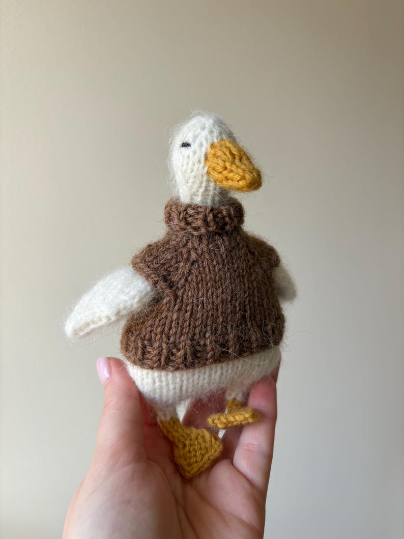May include: Hand-knitted duck toy with a white head, wings, and body, a yellow beak and feet, and a brown sweater. The duck is a soft, plush toy, perfect for children or as a decorative item.