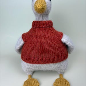 May include: A handmade knitted duck toy with a white body, gray wings, and a yellow beak and feet. The duck is wearing a red sweater. The toy is designed to be a soft and cuddly item.