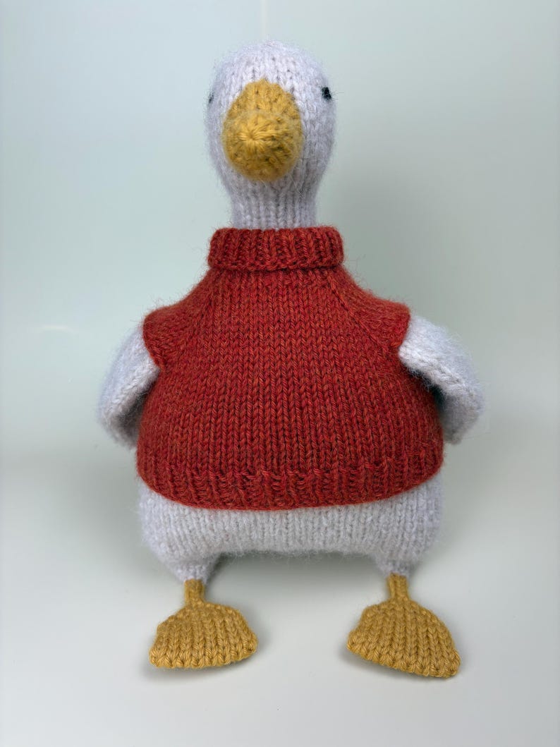 May include: A handmade knitted duck toy with a white body, gray wings, and a yellow beak and feet. The duck is wearing a red sweater. The toy is designed to be a soft and cuddly item.