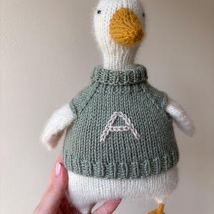 May include: A handmade knitted duck toy with a white body, a yellow beak and feet, and a green sweater. The sweater has a white embroidered letter "A" on the front. The duck has small black eyes.