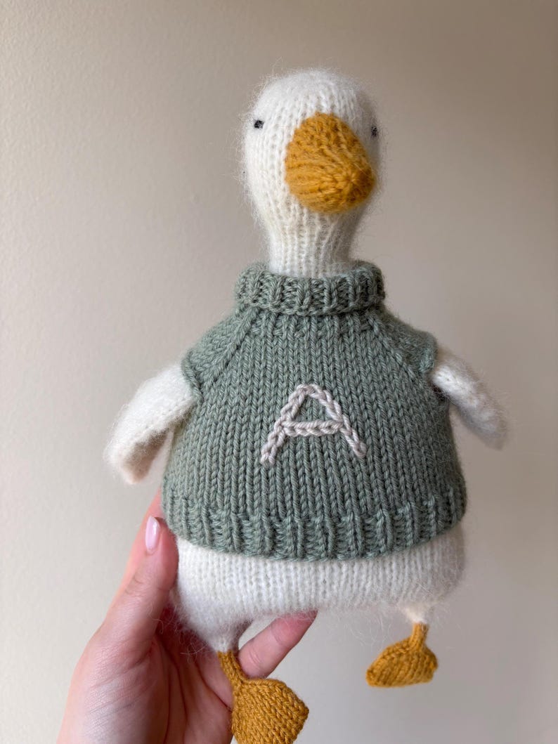 May include: A handmade knitted duck toy with a white body, a yellow beak and feet, and a green sweater. The sweater has a white embroidered letter "A" on the front. The duck has small black eyes.