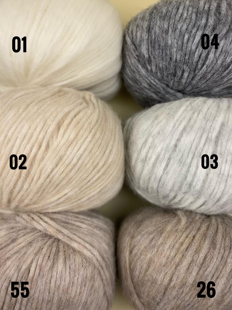 May include: Close-up of six skeins of yarn in various neutral colors. The yarn is labeled with numbers: 01, 02, 03, 04, 26, and 55. The colors range from off-white to light gray and beige. The yarn appears soft and fluffy.