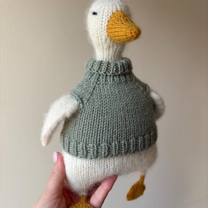 May include: A handmade knitted goose doll with a white body, a green sweater, and yellow beak and feet. The goose has a fluffy texture and a simple design, perfect for a child's toy or a decorative item.