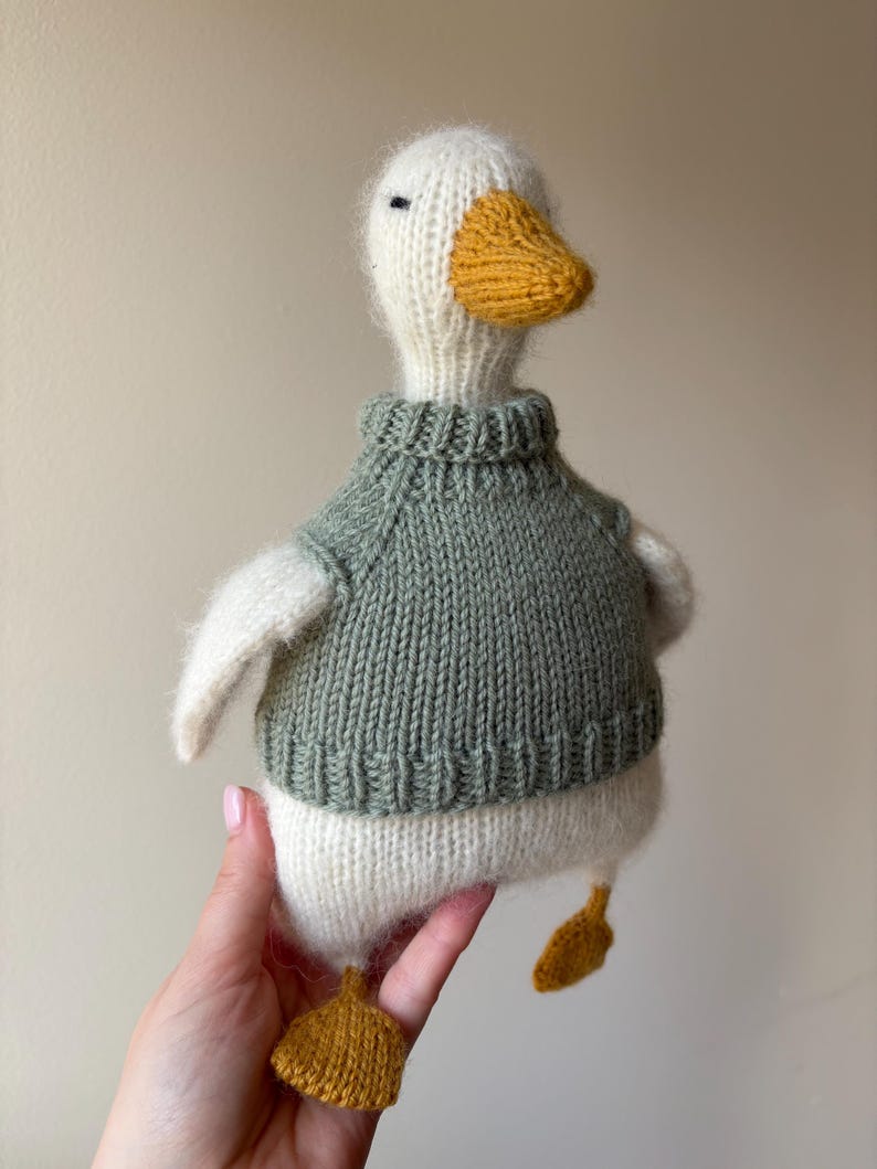 May include: A handmade knitted goose doll with a white body, a green sweater, and yellow beak and feet. The goose has a fluffy texture and a simple design, perfect for a child's toy or a decorative item.