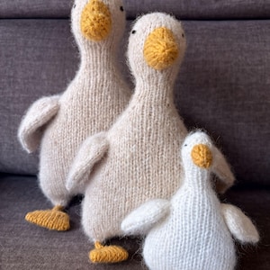 May include: Three knitted goose figurines in varying sizes. The geese are primarily off-white with yellow beaks and feet. The soft, textured appearance suggests a handmade craft item, perfect for home decor or a child's toy.