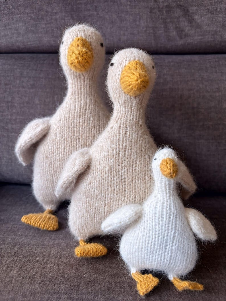 May include: Three knitted goose figurines in varying sizes. The geese are primarily off-white with yellow beaks and feet. The soft, textured appearance suggests a handmade craft item, perfect for home decor or a child's toy.
