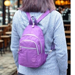 May include: A purple nylon backpack with a zippered main compartment and a smaller front pocket. The backpack has adjustable straps and a top handle. The zippers and trim are silver. The backpack is worn over the shoulder.