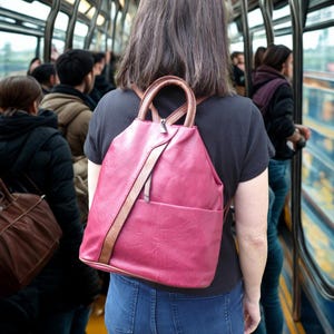 May include: A burgundy backpack with brown detailing is worn by a person in a black top and blue jeans. The backpack features a front pocket and a zip closure. The straps and handle are brown. The background shows other people on public transport.