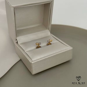 K&ouml;nnte beinhalten: Ein Paar Ohrringe aus Gold und wei&szlig;en Diamanten in einer wei&szlig;en Schachtel. Die Ohrringe sind klein und zart, mit einem einzelnen Diamanten in der Mitte jedes Ohrrings.