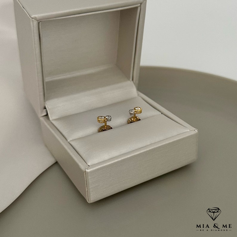 K&ouml;nnte beinhalten: Ein Paar Ohrringe aus Gold und wei&szlig;en Diamanten in einer wei&szlig;en Schachtel. Die Ohrringe sind klein und zart, mit einem einzelnen Diamanten in der Mitte jedes Ohrrings.