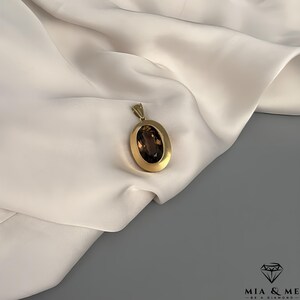 May include: A gold pendant with an oval, brown gemstone. The pendant is set in a gold frame and hangs from a small loop. The background is a white, draped fabric.