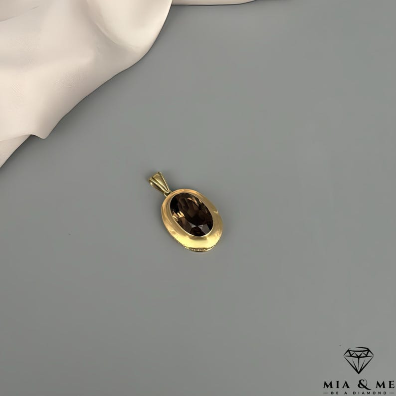 May include: A gold pendant with an oval, smoky quartz gemstone. The pendant has a simple, elegant design with a gold bail. The gemstone is a rich, dark brown color, set in a gold bezel.