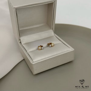 K&ouml;nnte beinhalten: Ein Paar kleine Diamant-Ohrringe in einer wei&szlig;en und goldenen Schmuckschatulle. Die Ohrringe bestehen aus zwei kleinen runden Diamanten, einem wei&szlig;en und einem goldenen.