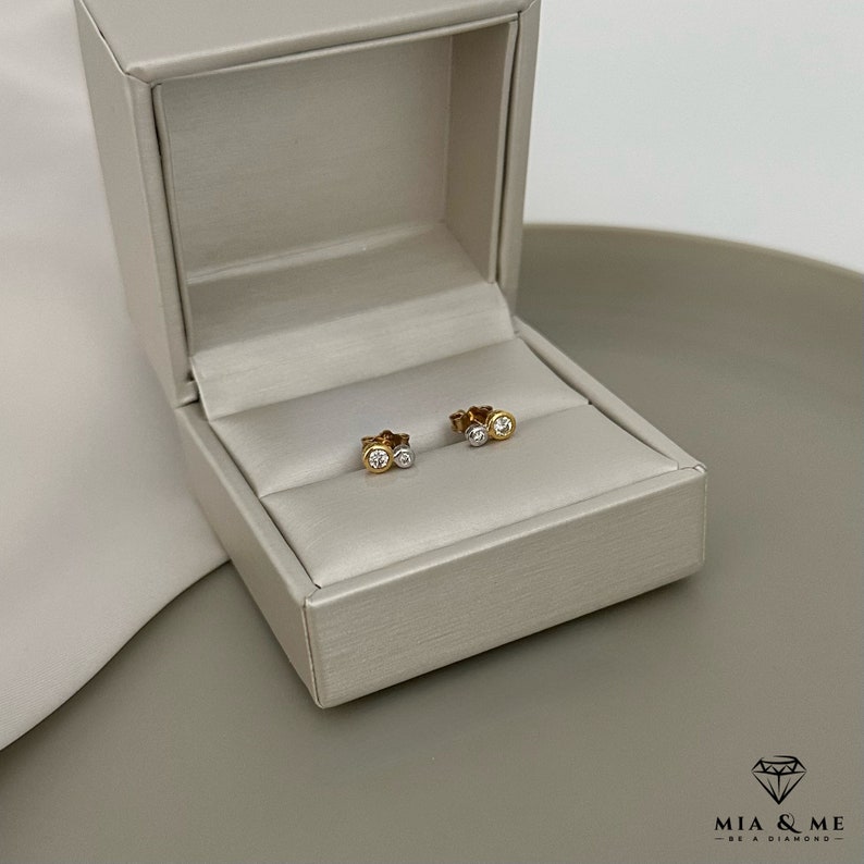 K&ouml;nnte beinhalten: Ein Paar kleine Diamant-Ohrringe in einer wei&szlig;en und goldenen Schmuckschatulle. Die Ohrringe bestehen aus zwei kleinen runden Diamanten, einem wei&szlig;en und einem goldenen.