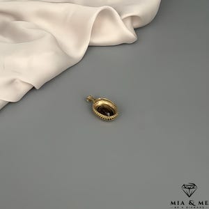 May include: A gold-colored pendant with a dark, oval-shaped stone at its center. The pendant has a decorative, beaded border and a small loop for a chain. The background includes a draped, off-white fabric and a gray surface.