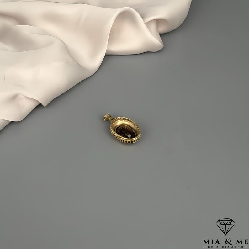 May include: A gold-colored pendant with a dark, oval-shaped stone at its center. The pendant has a decorative, beaded border and a small loop for a chain. The background includes a draped, off-white fabric and a gray surface.