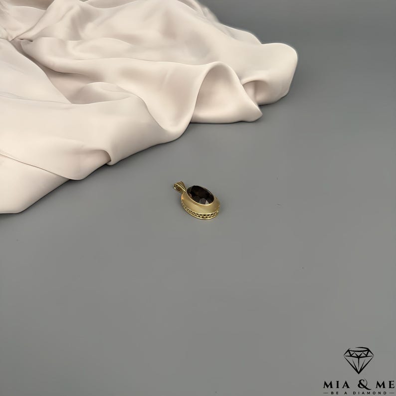 May include: A gold pendant with an oval, dark gemstone. The pendant has a textured, decorative border. The background is a light gray surface with a cream-colored fabric draped in the upper left corner. The pendant is a piece of jewelry.