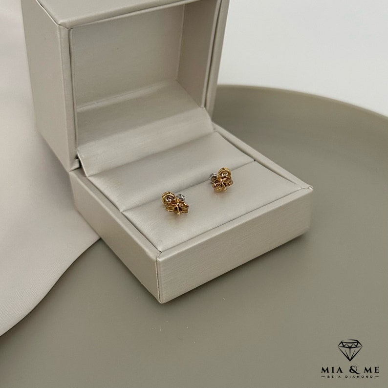 K&ouml;nnte beinhalten: Ein Paar goldene und silberne Ohrringe in Blumenform in einer wei&szlig;en Schachtel. Die Ohrringe sind klein und zart.
