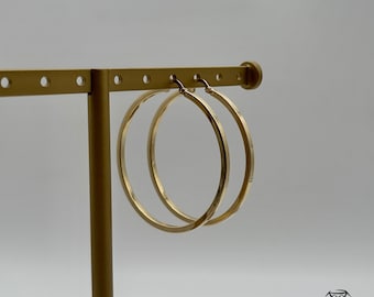 Könnte beinhalten: Ein Paar goldene Creolen, die an einem Ausstellungsständer hängen. Die Ohrringe sind schlicht und elegant, mit einer glatten, polierten Oberfläche.