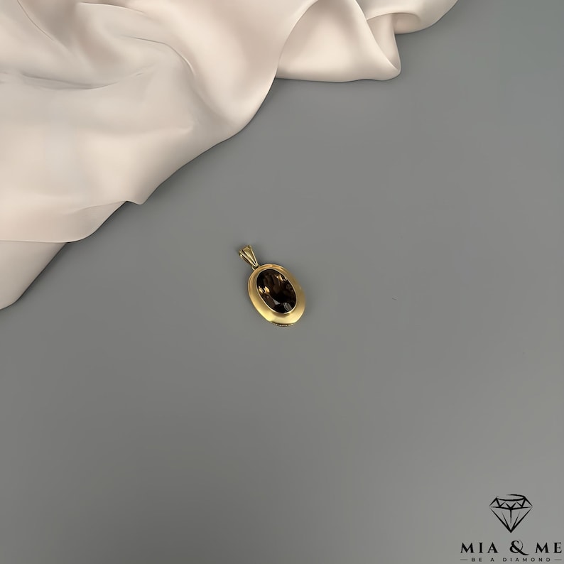 May include: A gold pendant with an oval, dark brown gemstone. The pendant has a simple, elegant design and is set against a gray background with a cream-colored fabric in the upper left corner. The pendant is a piece of jewelry.