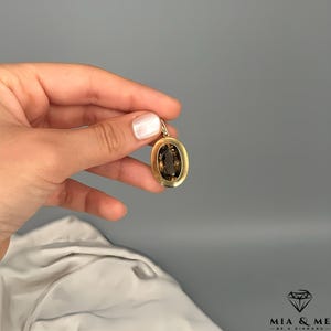 May include: A gold pendant with an oval, smoky-brown gemstone. The pendant is held against a gray background. The gemstone is set in a gold frame. The pendant is a piece of jewelry.