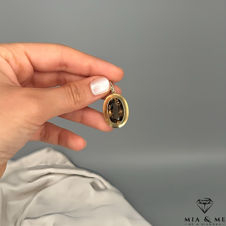 May include: A gold pendant with an oval, smoky-brown gemstone. The pendant is held against a gray background. The gemstone is set in a gold frame. The pendant is a piece of jewelry.