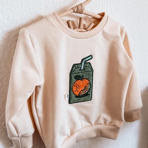 Könnte beinhalten: Ein hellbeiges Langarm-Sweatshirt mit einem zentralen gestickten Aufnäher einer grünen Saftpackung mit Orangenscheiben-Grafik und blauem Strohhalm. Das Kleidungsstück hat einen Rundhalsausschnitt sowie gerippte Bündchen an Ärmeln und Saum.